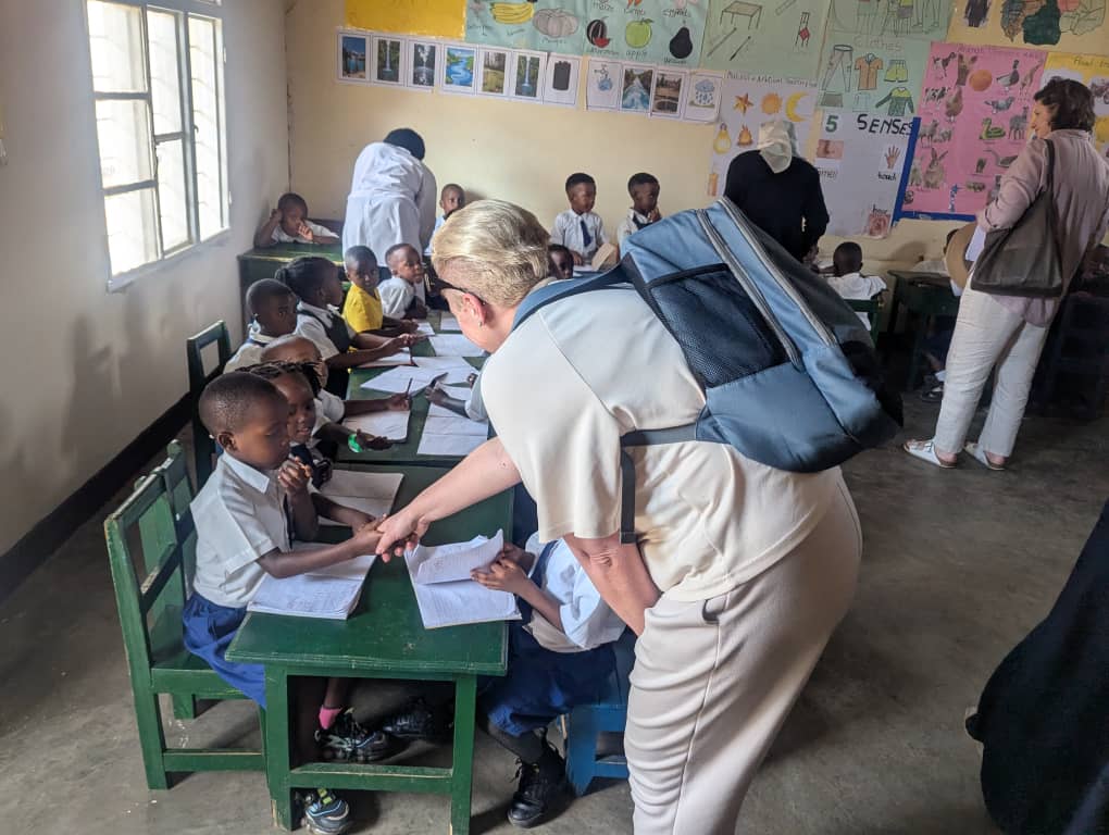 Visitors from Rosa Mystica interact with young students in a classroom, leaning over desks to look at their notebooks while children in school uniforms sit together and work on lessons. The classroom walls are decorated with colorful educational posters, and teachers assist students in the background