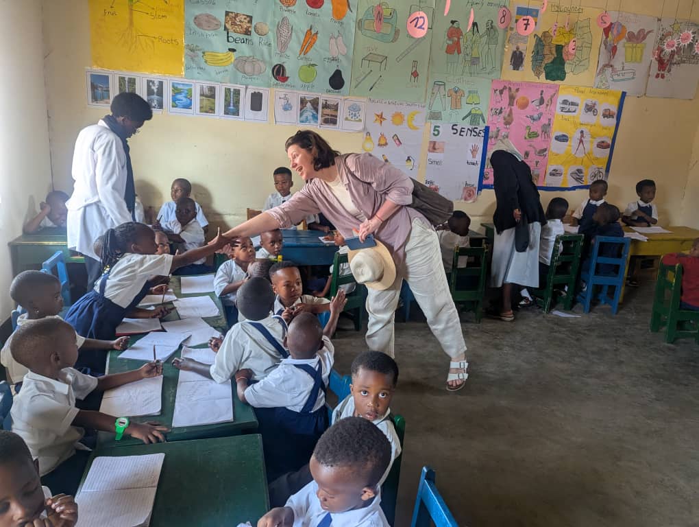 A visitor greets and interacts with young students seated at desks in a colorful classroom while teachers and a nun assist nearby, during a visit to Rosa Mystica school.