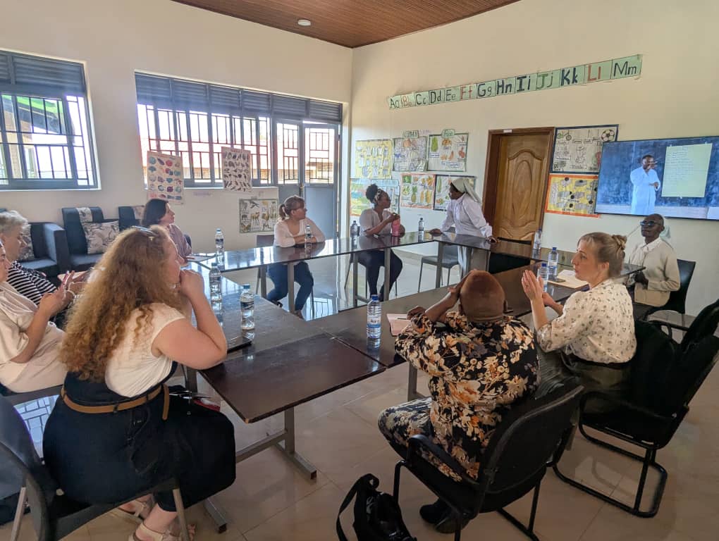 Visitors sit around tables in a classroom discussing with a nun and staff member while viewing a presentation, with educational posters and the alphabet displayed on the walls during a Rosa Mystica visit.