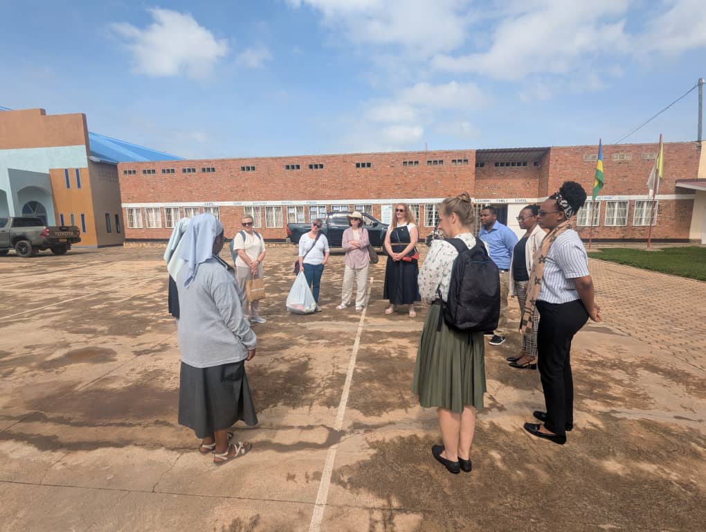 A group of visitors stands in a circle in a school courtyard while a nun speaks to them, with a brick school building and flags in the background during a GS Rosa Mystica visit.